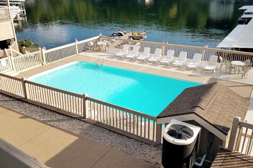 A spacious outdoor swimming pool is surrounded by a wooden deck, with several lounge chairs placed around it. The clear blue water reflects the surrounding landscape, while docks for boats are visible on the serene lake in the background.