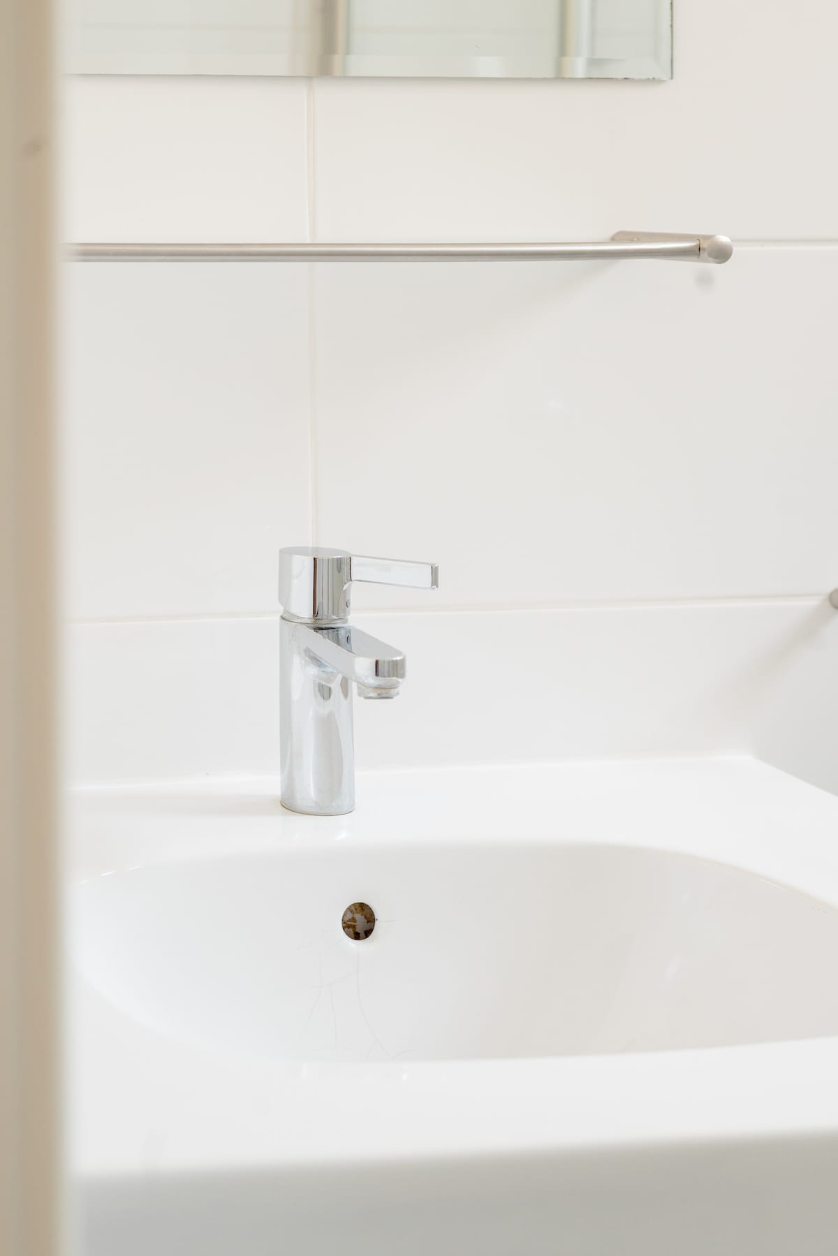 A modern bathroom sink is equipped with a chrome faucet set against a clean, white tiled wall. The sink features a smooth, rounded basin, and a drain is visible at the center, contributing to a minimalist aesthetic.