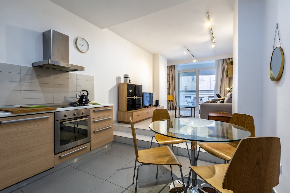 A modern kitchen and dining area features light wood cabinetry and a circular glass table surrounded by four wooden chairs. Natural light from the adjacent living area enhances the neutral decor, with a large window providing views of the outdoor terrace.