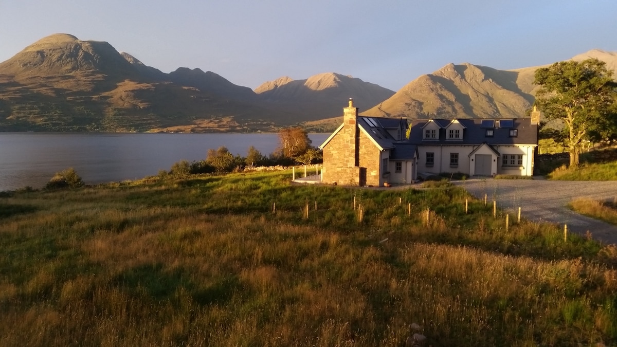 A stone cottage is positioned beside a tranquil loch, surrounded by lush grass and highland scenery. Majestic mountains rise in the background, bathed in golden light from the setting sun. A gravel path leads to the cottage, enhancing its secluded and natural setting.
