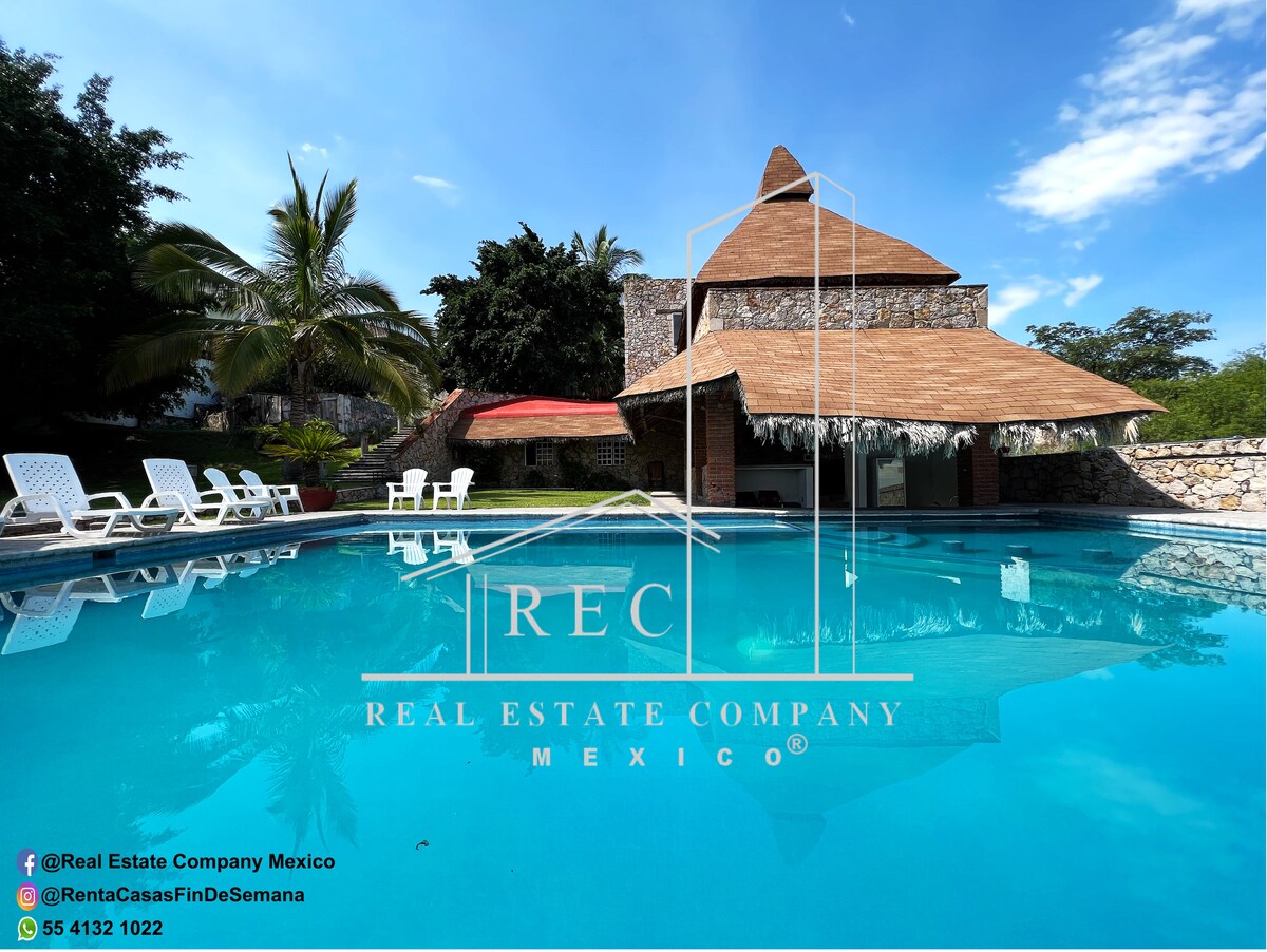 A large swimming pool is surrounded by palm trees and sun loungers, creating a relaxing space. A palapa-style structure provides shade nearby. The bright blue sky reflects off the pool's surface, enhancing the serene atmosphere of the outdoor area.