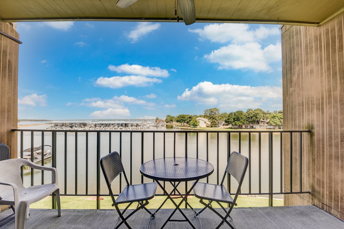 A private balcony offers unobstructed views of the water and marina. A round table with two chairs is positioned on the balcony, providing a space for relaxation. The background features green trees and blue skies, enhancing the serene lakeside atmosphere.