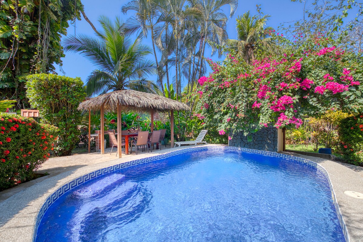 A vibrant outdoor area showcases a clear swimming pool surrounded by lush tropical greenery and flowering plants. A thatched-roof rancho is seen nearby, furnished with a dining table and several chairs, inviting relaxation in a picturesque garden setting.