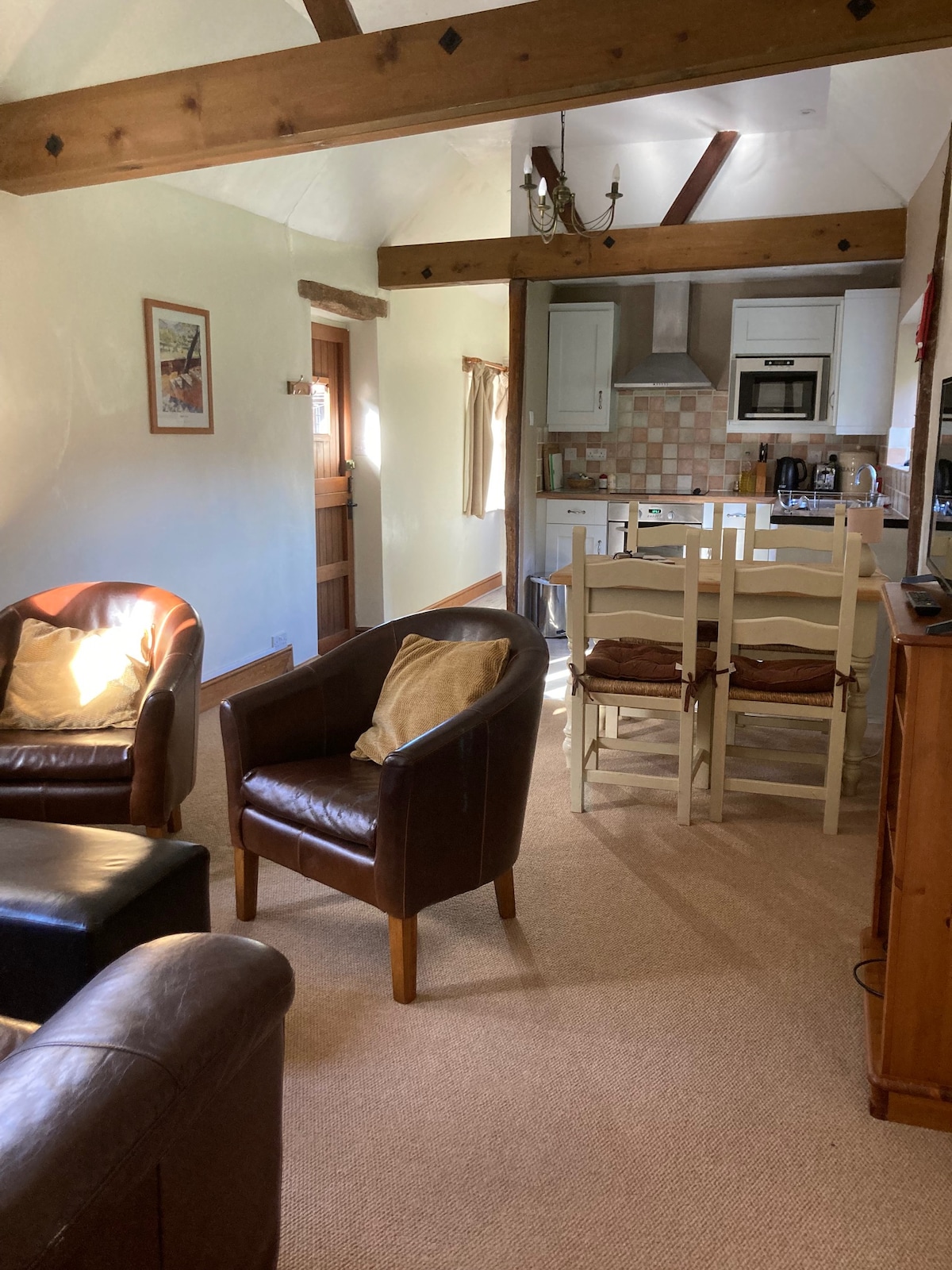 An inviting living area features two leather armchairs and a cozy sofa, providing comfortable seating. A central dining area with a table and four chairs is visible. Warm light filters through a nearby window, highlighting the rustic beams and soft carpeting.