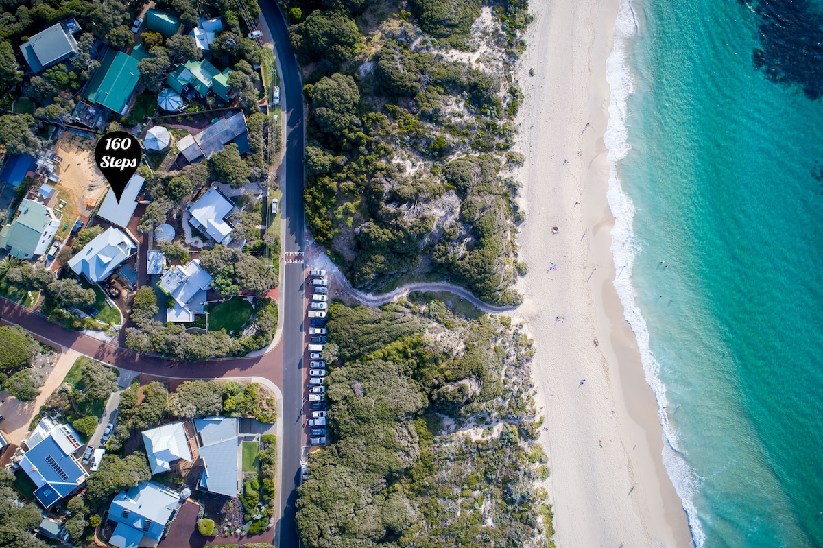 An aerial view captures 160 Steps nestled among a cluster of homes adjacent to Yallingup Beach. The sandy shoreline meets the turquoise waters, with pathways visible along the beach. The surrounding greenery adds a touch of nature to the vibrant coastal setting.