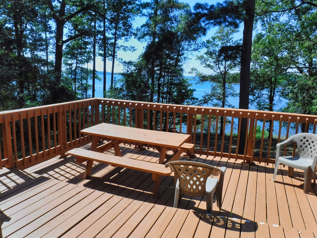 A wooden deck is shown, featuring a picnic table and several plastic chairs. The view beyond includes a serene lakeside framed by trees, with sunlight casting soft shadows across the deck.