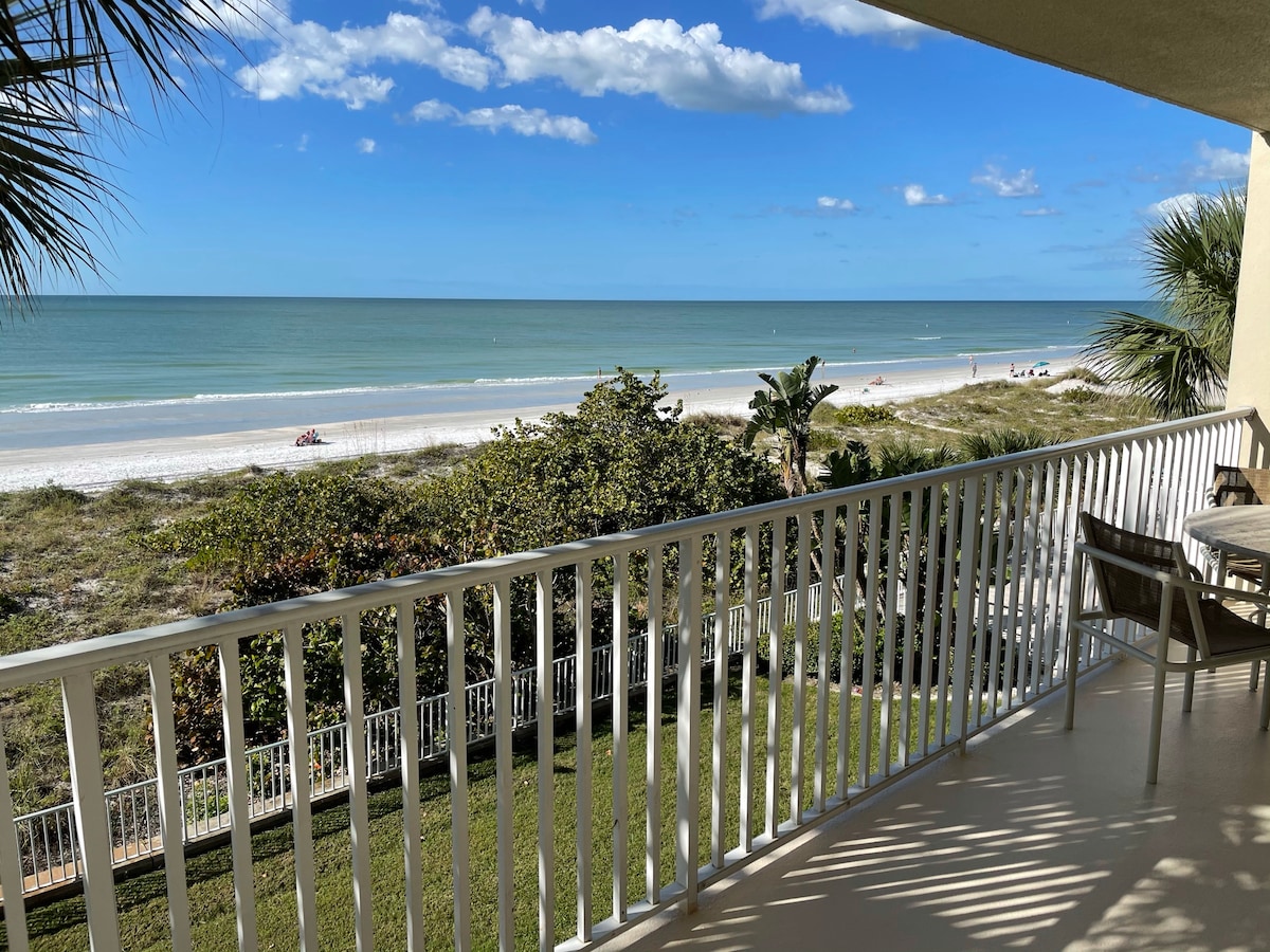 A spacious balcony offers direct views of Indian Rocks Beach and the Gulf of Mexico. Lush greenery frames the scene, while soft waves can be seen lapping at the shore under a clear blue sky.
