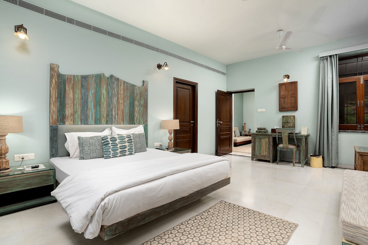 A spacious guest room features a large bed with a rustic wooden headboard. Natural light floods the room through a window, with soft blue walls complementing wooden furniture. A small work desk and stylish lamps provide functionality, while a patterned rug adds a touch of comfort.