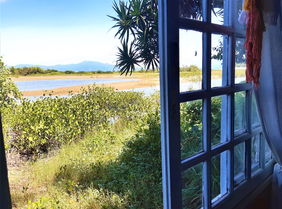 A view from a window reveals lush greenery and a tranquil lagoon. The landscape features sandy stretches and distant mountains, under a clear blue sky. Sunlight filters through the window, providing a bright and inviting atmosphere.