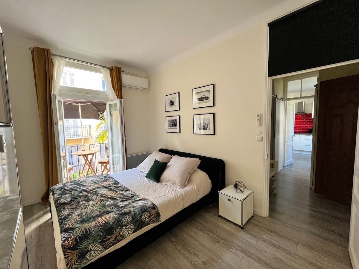A bright bedroom is presented, featuring a spacious bed adorned with patterned bedding. Light streams through open doors that lead to a small balcony. Artwork graces the walls, and a compact side table is positioned beside the bed.