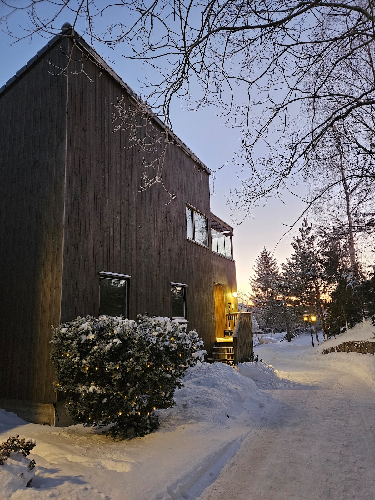 The exterior of the house is shown at dusk, framed by a snowy pathway leading to the entrance. A wooden facade complements the winter setting, while soft lighting illuminates the entrance. Tall trees and a subtle sunset provide a serene backdrop.