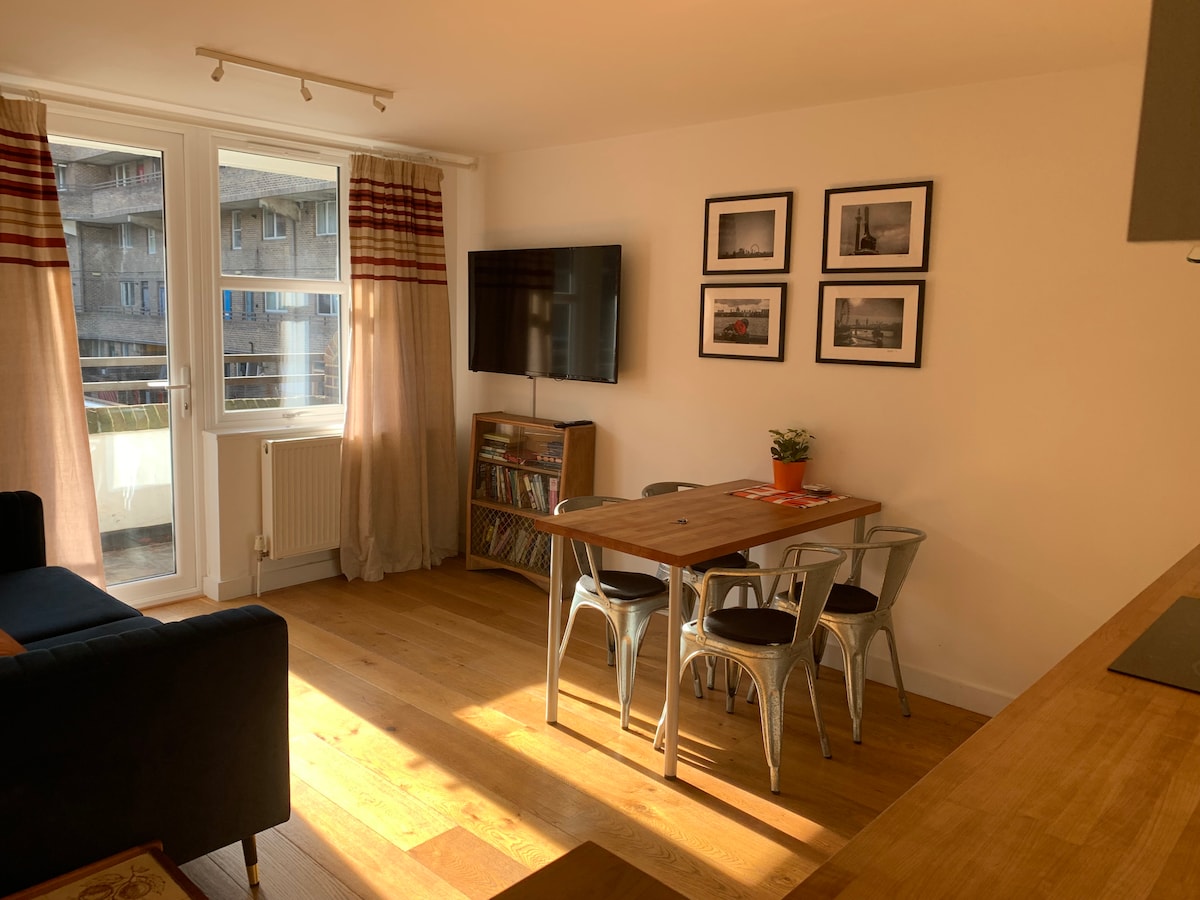 A bright living area features a comfortable seating arrangement and a wooden dining table surrounded by metal chairs. Natural light filters through large windows, accentuated by light curtains. Black-and-white photographs adorn the walls, complementing a media console with colorful records.