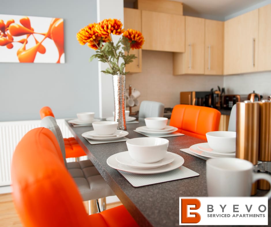 A modern dining area is presented, featuring a long table set with white bowls and orange chairs. A vase filled with vibrant autumn flowers adds a decorative touch. The open kitchen space is visible in the background, equipped with sleek cabinetry and appliances.