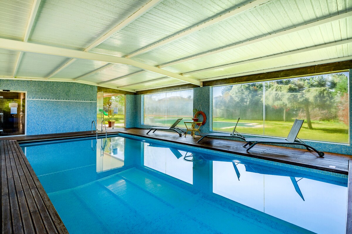 An indoor swimming pool is enclosed by large glass windows, allowing natural light to illuminate the space. Lounge chairs are positioned around the pool on a wooden deck, creating a relaxing environment. The walls feature a calming blue design, complementing the tranquil water.