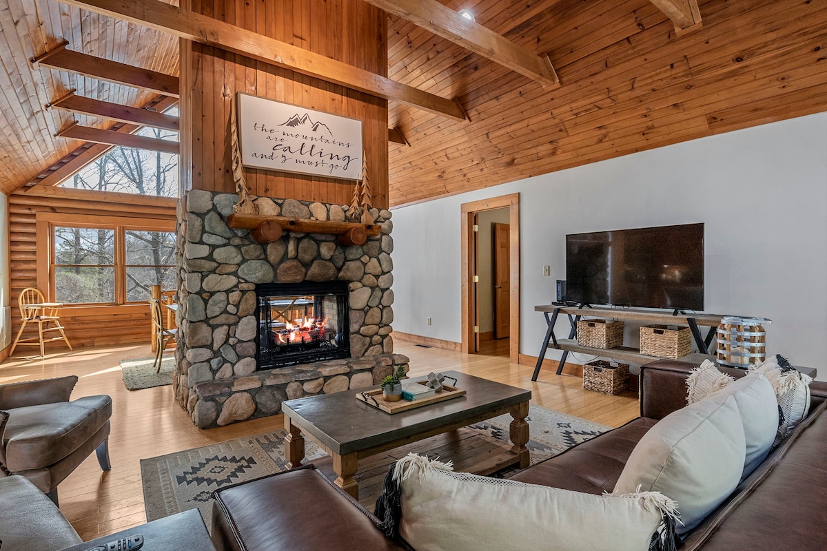 A comfortable living area features a stone fireplace with a cozy fire. Wooden beams and walls create a warm ambience. A large television is positioned on a stand across from a sectional sofa, with a coffee table in the center. Natural light floods in through expansive windows.