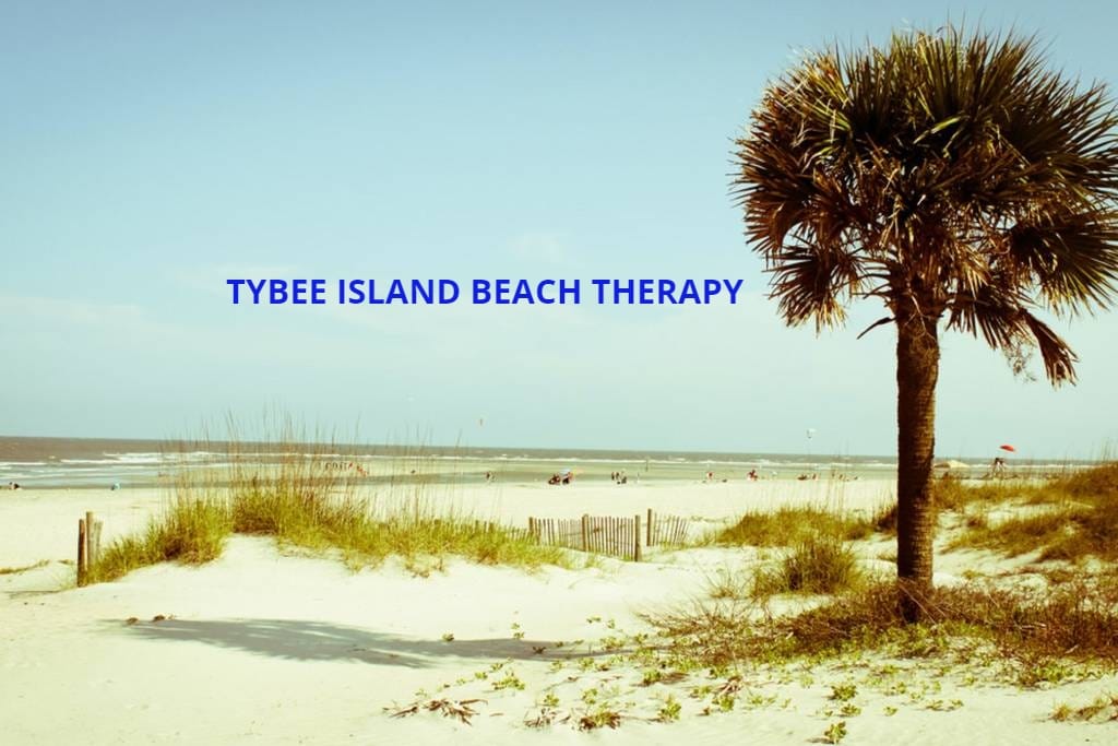 A sandy beach is seen with gentle waves in the distance. Lush greenery and tall grasses frame the foreground, while a palm tree adds a tropical touch. The sky is bright with a few clouds, suggesting a warm, inviting atmosphere.
