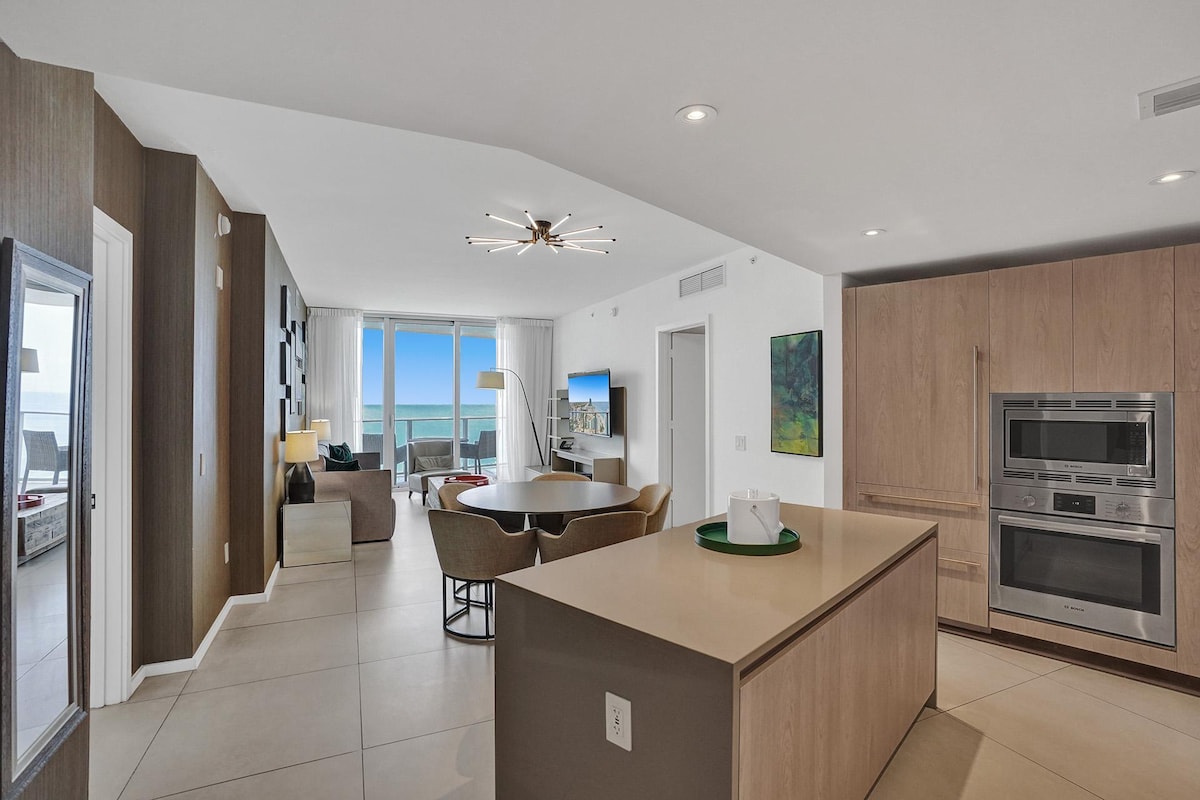 An open-concept kitchen and living area features modern design elements, including wooden cabinetry and sleek appliances. Large windows provide expansive views of the ocean, creating a bright atmosphere. A round dining table with stylish chairs complements the space, while a comfortable seating area invites relaxation.