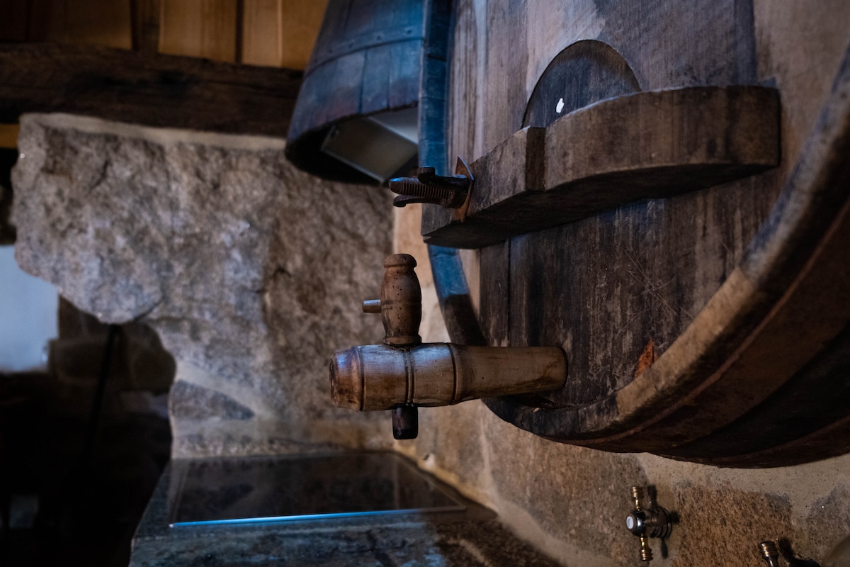 A rustic wooden barrel is mounted on a stone wall, featuring a wooden tap. The grain of the wood is visible and hints at its age. A stone countertop below complements the natural materials used throughout the space.