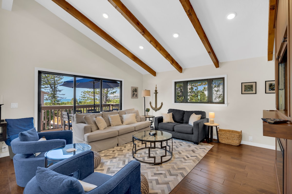 A bright living area features vaulted beamed ceilings and large windows offering views of the outdoors. Comfortable seating includes a beige sofa and blue accent chairs, arranged around a glass coffee table on a geometric patterned rug. Natural light floods the space, giving it an airy feel.