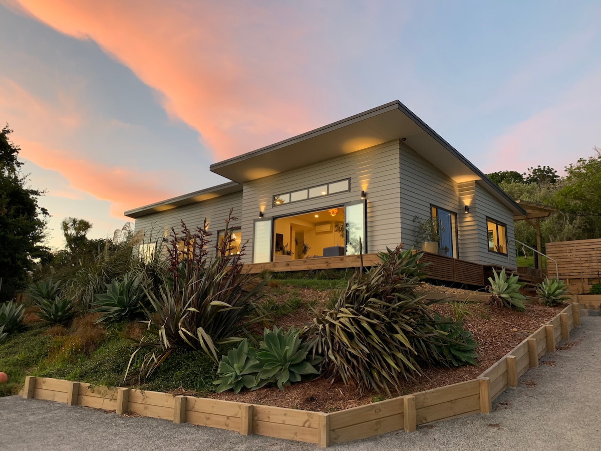 The exterior of the modern home is showcased against a colorful sunset sky. Lush greenery, including various plants and shrubs, surrounds the property. Large windows invite natural light into the space, highlighting the inviting design of the home.