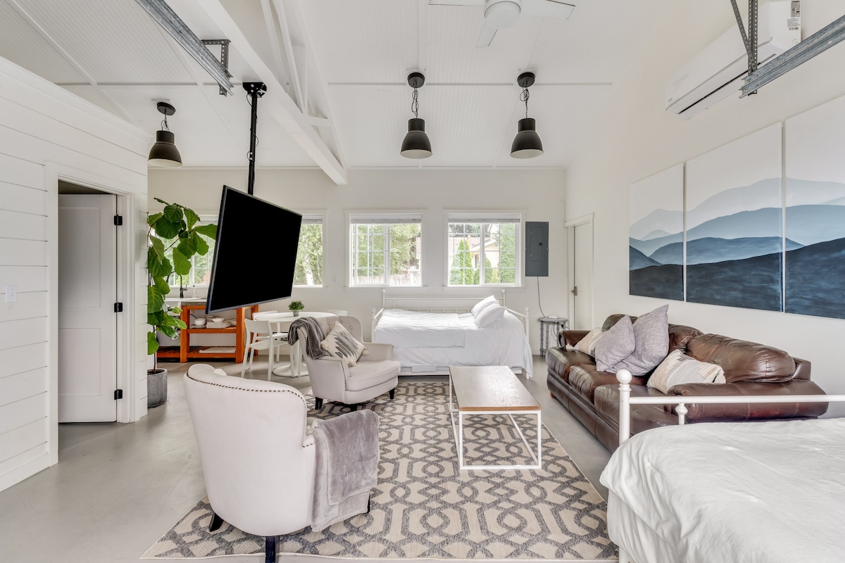 An open-concept studio space features two separate sleeping areas with full beds and a twin trundle bed. A large window allows for ample natural light, showcasing a comfortable seating arrangement with a leather sofa and a cozy armchair. An HDTV is mounted for entertainment.