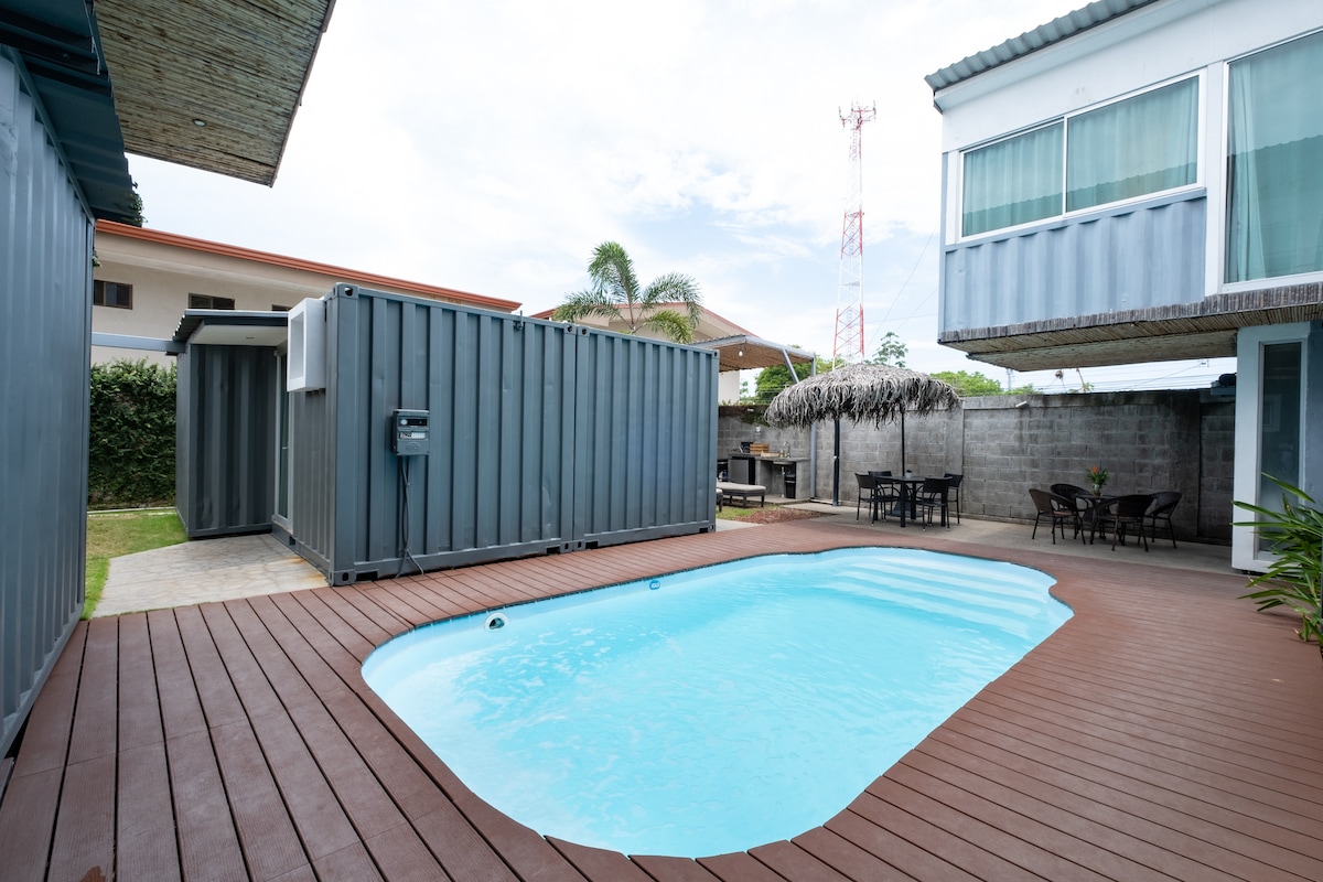 An inviting swimming pool is surrounded by a wooden deck, providing a relaxing space. Nearby, a thatched-roof structure and outdoor seating area offer shaded options for lounging. The area is framed by eco-friendly container structures and greenery, creating a serene environment.