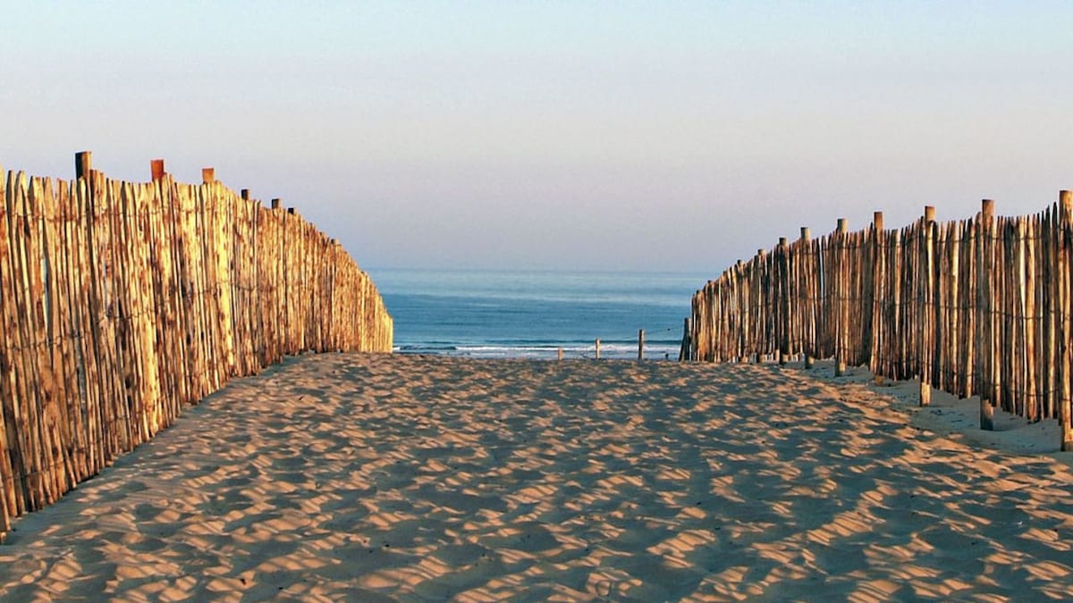 A sandy pathway leads between wooden fences towards the ocean. Gentle waves can be seen in the distance, creating a serene coastal view. The horizon features soft colors of a setting sun, enhancing the peaceful ambiance of the beach entrance.
