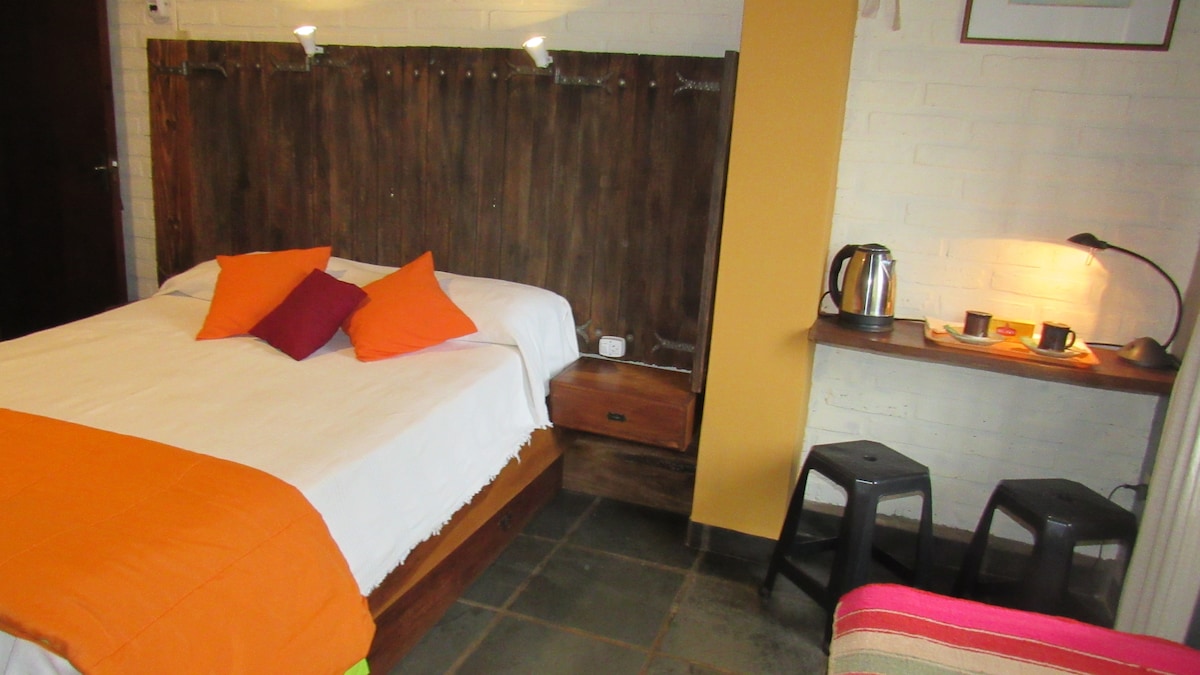 A comfortable sleeping area features a spacious bed with a white duvet and vibrant orange and purple decorative pillows. A wooden headboard creates a warm backdrop, while a side table holds a kettle and drinks. Soft lighting is provided by a desk lamp and wall fixtures.