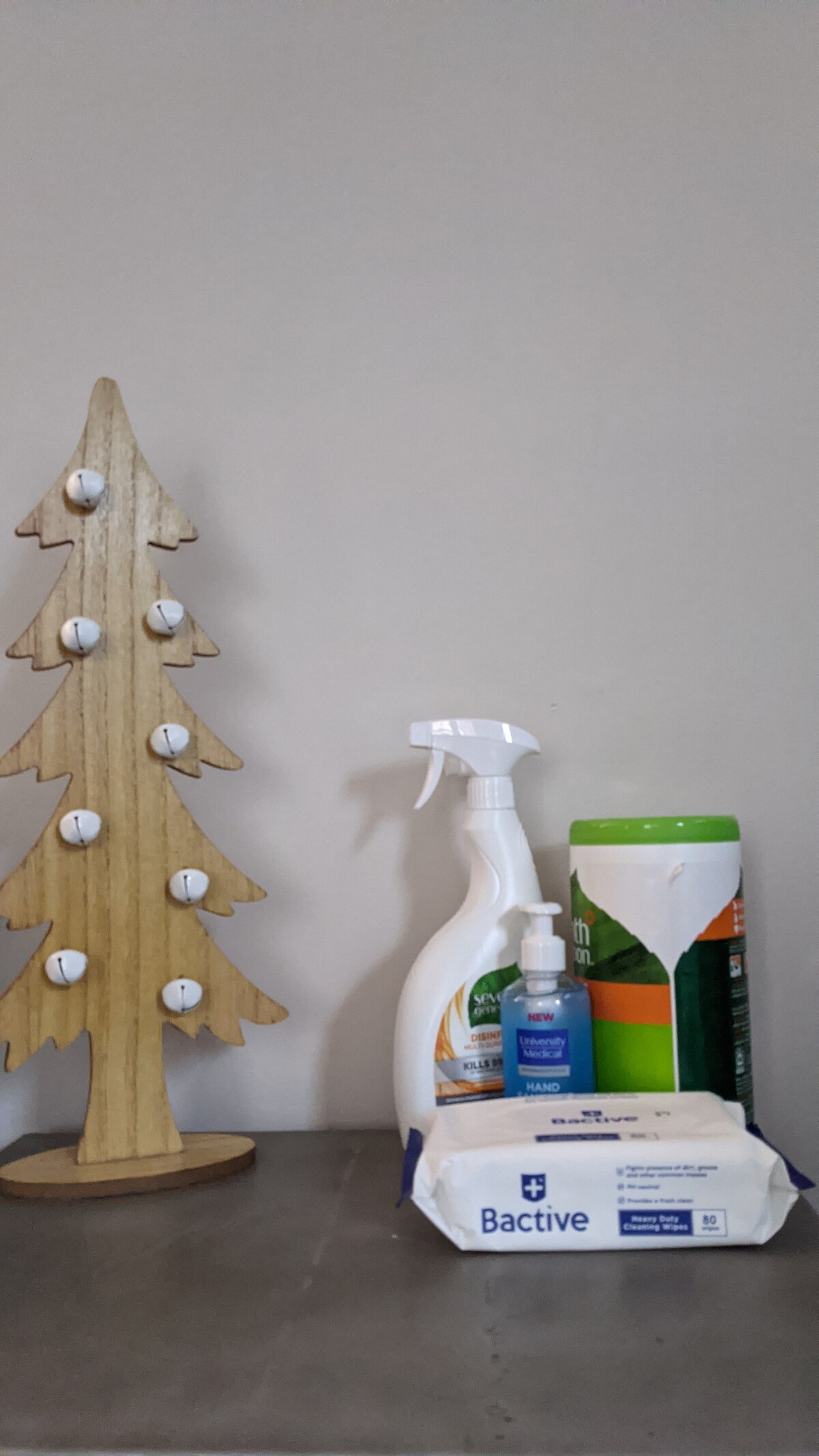 A wooden tree-shaped shelf is displayed against a light wall. Positioned on a flat surface, cleaning supplies including a spray bottle, hand sanitizer, and a pack of wipes are arranged next to a green container, showcasing practicality for maintaining cleanliness.
