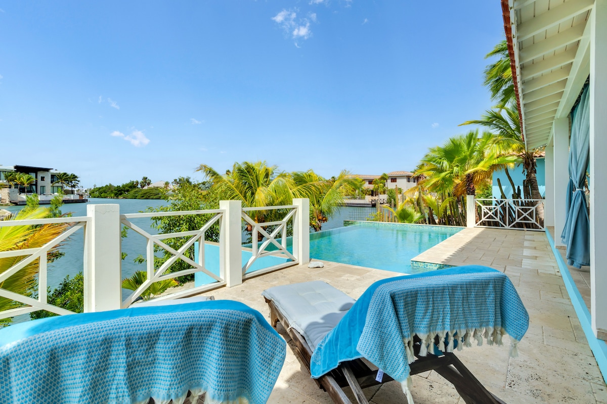 A spacious terrace features two loungers draped with blue towels, overlooking an infinity pool. Palm trees surround the area, providing a tropical ambiance. The lagoon is visible in the background under a clear blue sky, enhancing the serene outdoor setting.