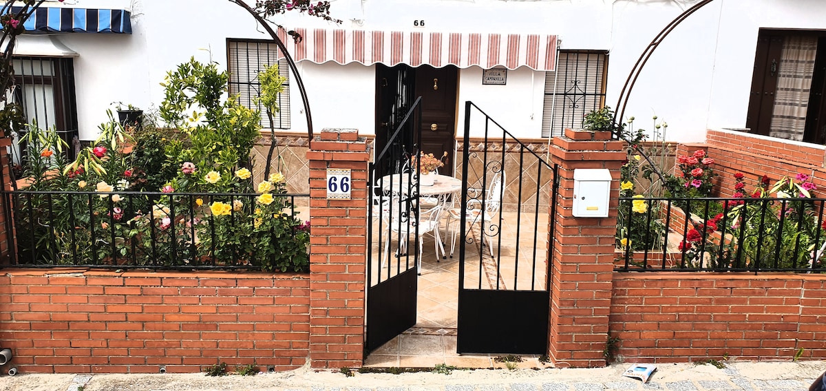 The entrance features a black wrought iron gate opening into a flower-filled garden with vibrant roses and assorted plants. A charming patio with white furniture can be seen beyond the gate, framed by the arched entryway and decorative brick wall.