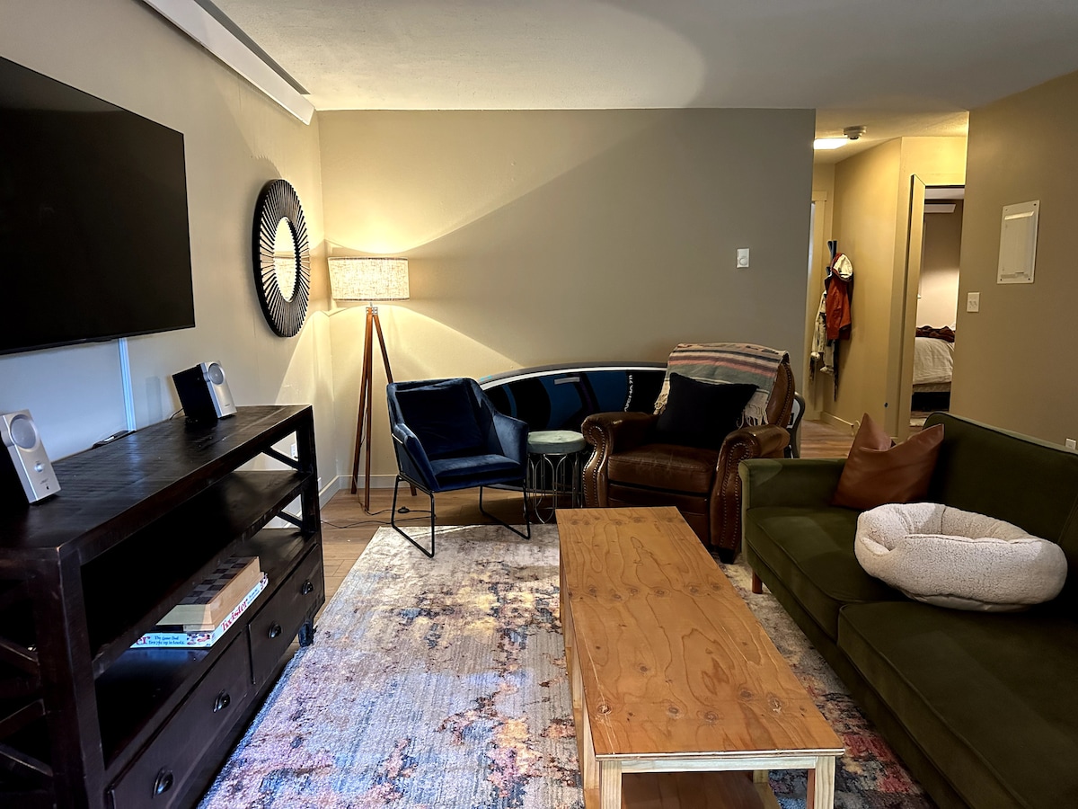 A living area features a comfortable arrangement of seating, including a leather armchair and a green sofa. A wooden coffee table sits at the center, and a large television is mounted on the wall. Soft lighting is provided by a floor lamp and a wall mirror adds depth.