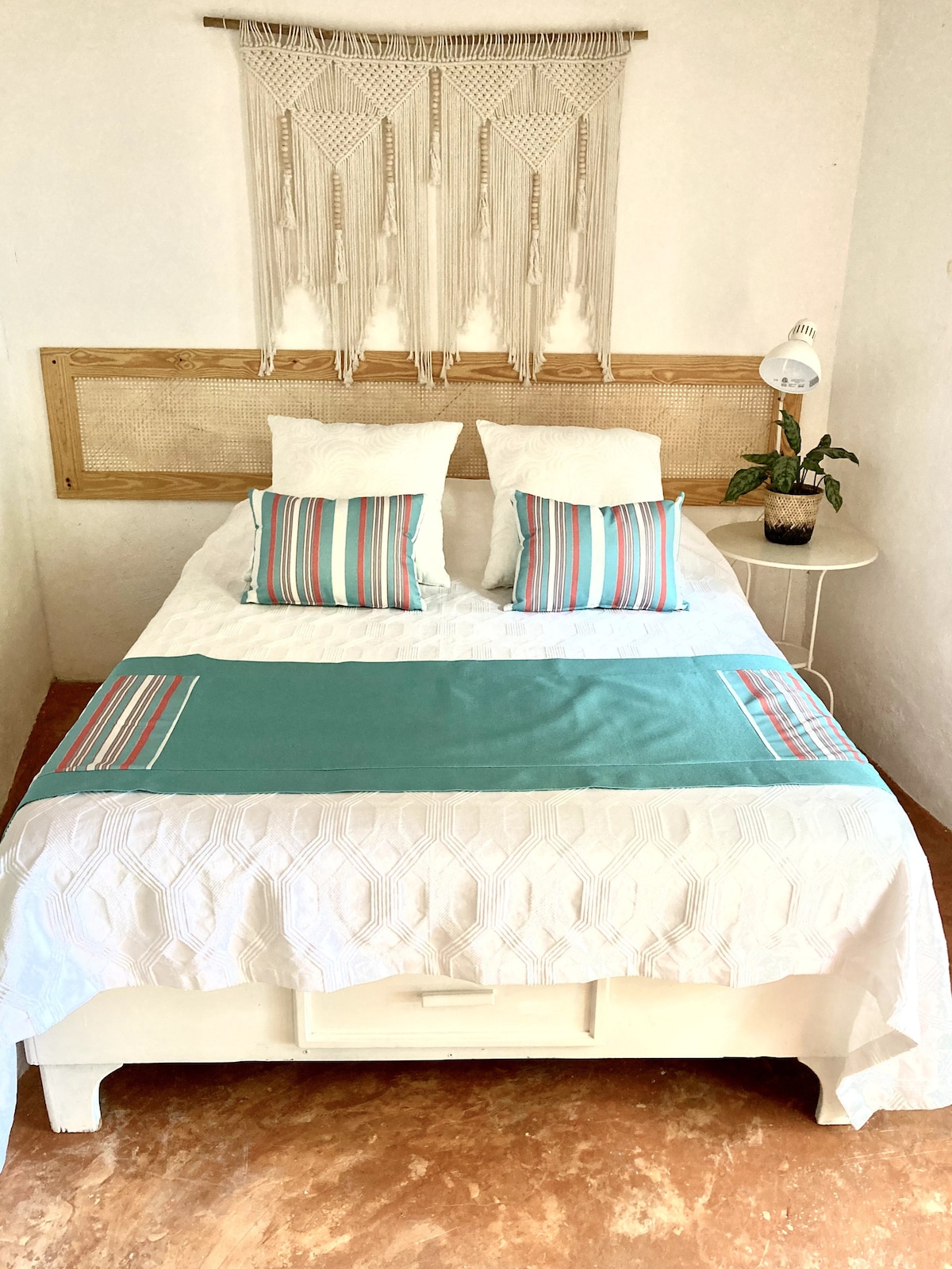 A cozy sleeping area features a spacious bed with striped accent pillows and a textured white duvet. A macramé wall hanging adds character above the bed. A small side table with a lamp and a potted plant contributes to the overall design.