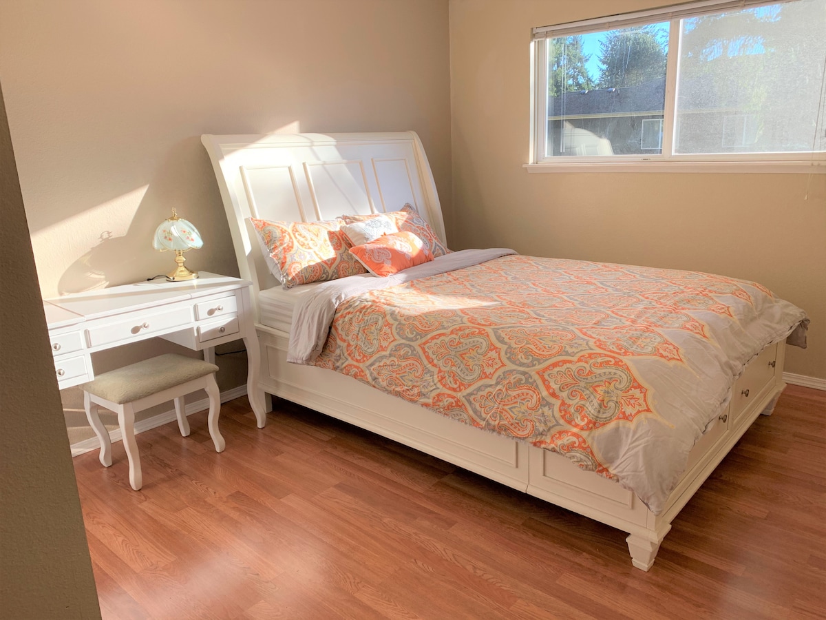 A well-lit bedroom features a spacious bed with a patterned comforter and coordinating pillows. A white nightstand holds a decorative lamp, and a small stool is positioned nearby. Natural light enters through a window, illuminating warm wood flooring.