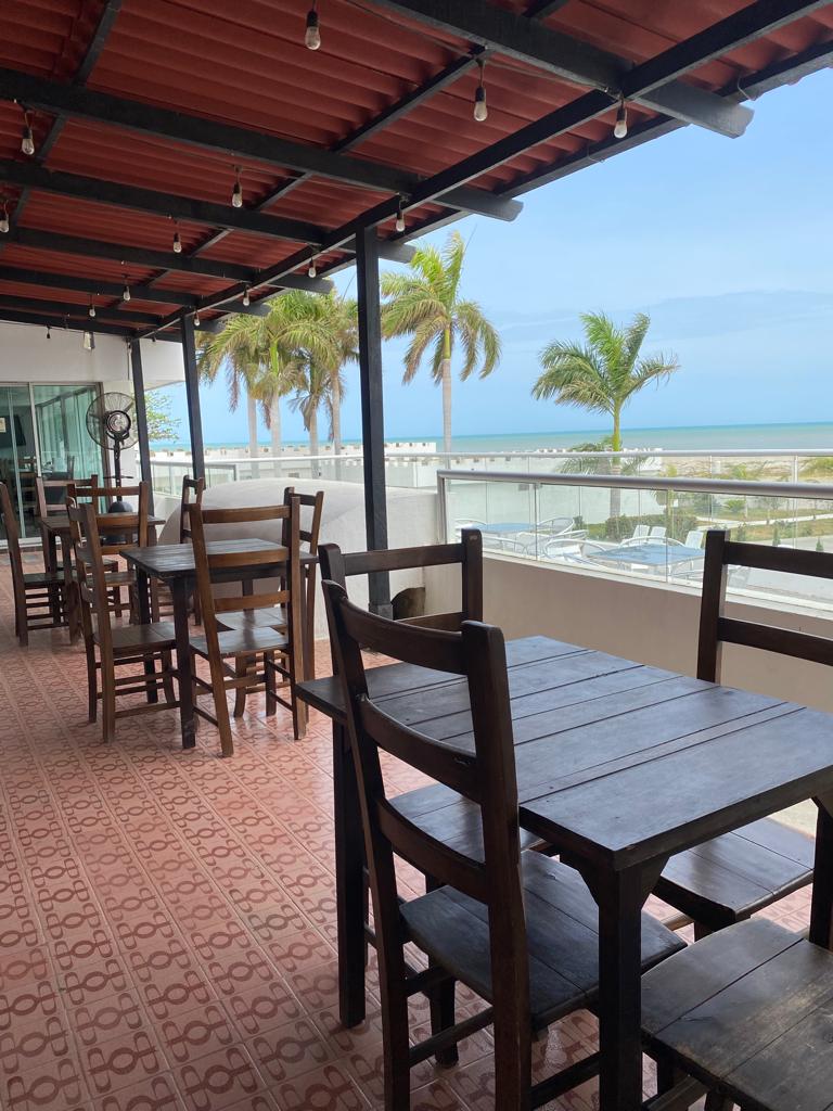 An open terrace is featured with several wooden tables and chairs, offering a clear view of the ocean and palm trees in the background. The roof is supported by dark beams, and natural light illuminates the space, providing a relaxed atmosphere.