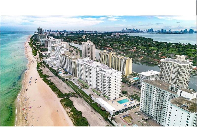 An aerial view captures the coastline, showcasing the sandy beach and clear turquoise waters. Several high-rise buildings line the shore, while the Miami skyline is visible in the distance. The beach is dotted with umbrellas and sunbathers, creating a vibrant seaside atmosphere.