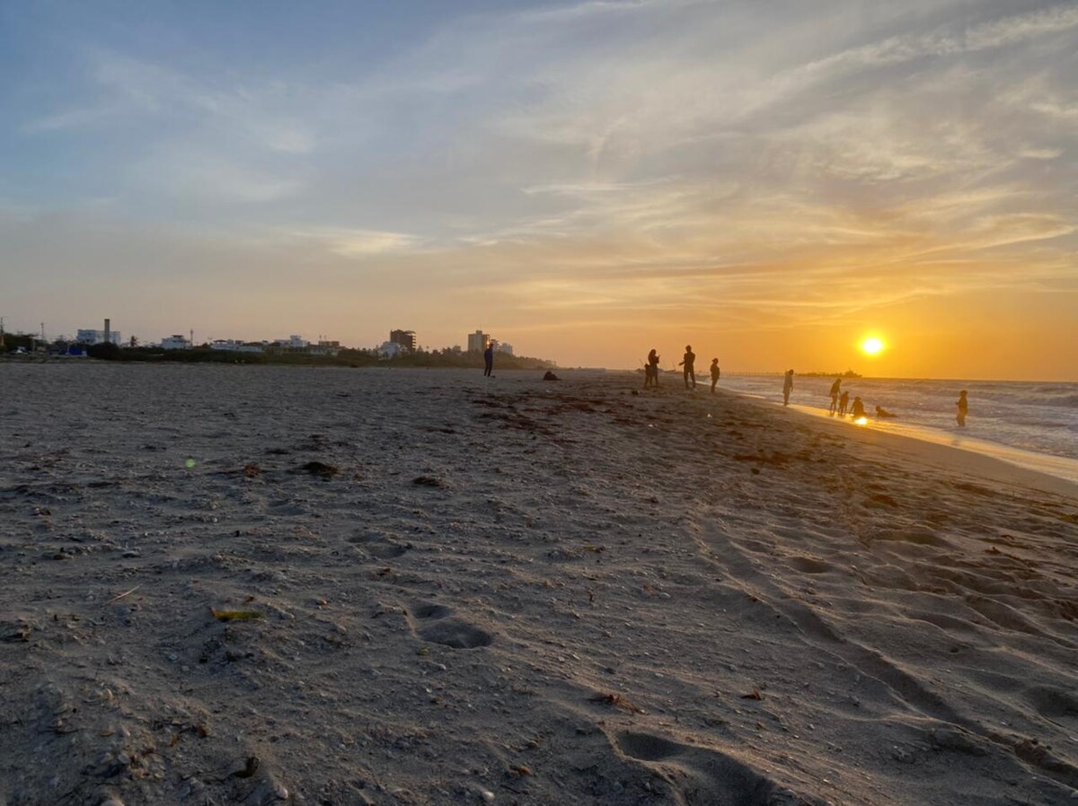 The sandy beach is shown at sunset, with gentle waves lapping at the shore. Silhouettes of people can be seen walking along the water's edge, while the sun casts a warm glow over the scene, creating a serene atmosphere.