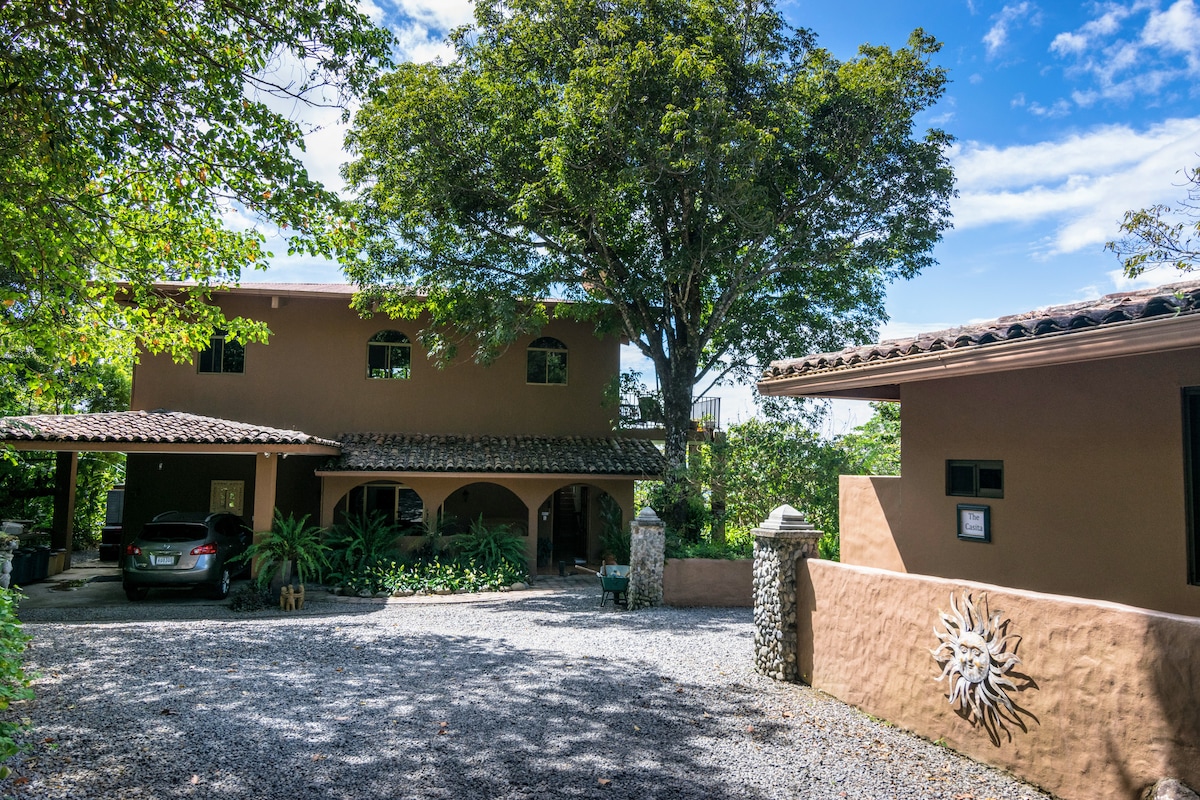 The exterior of The Hacienda is presented, featuring a two-story structure surrounded by lush greenery. A spacious gravel driveway accommodates a vehicle, while the prominent tree offers shade. The charming entrance showcases a decorative sun motif, enhancing the inviting feel of the property.