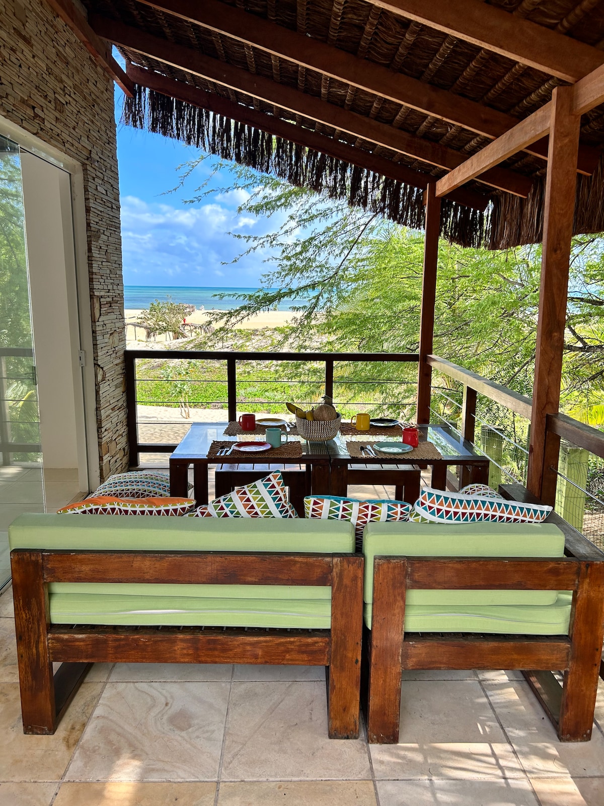 A shaded terrace is furnished with a wooden table and two comfortable seating areas, offering a relaxing space for meals or gatherings. Bright decorative cushions add color to the seating. The ocean can be seen in the background, surrounded by lush greenery.