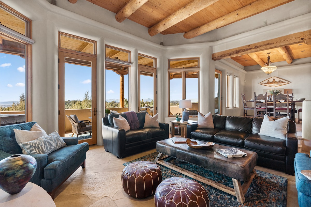 A spacious living area showcases large windows offering views of the surrounding landscape. Elegant leather sofas and a textured coffee table create a comfortable seating arrangement. A chandelier hangs above a dining area in the background, with stylish decor enhancing the inviting atmosphere.