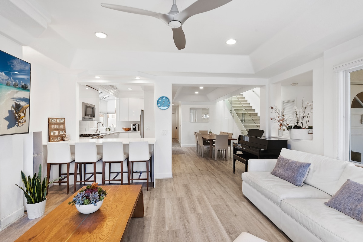 This spacious living area features a light wood floor and modern furnishings. A white sectional sofa is complemented by a wooden coffee table and a tall plant. The open-concept design includes a sleek kitchen with bar seating and a dining table visible in the background.
