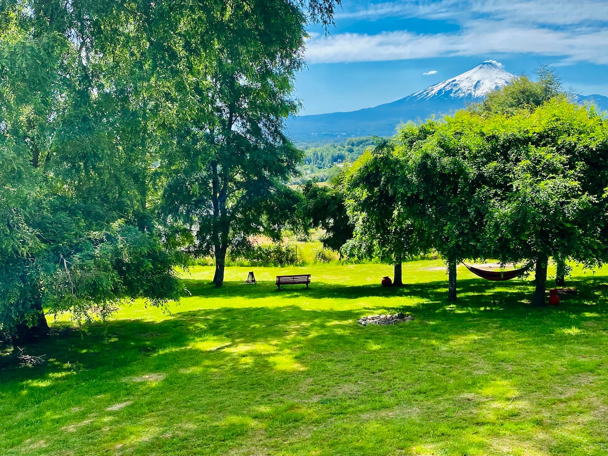 A serene outdoor space is displayed, featuring lush green grass and mature trees.  A hammock is suspended between two trees, and a wooden bench is positioned nearby. The view of the snow-capped Villarrica volcano is prominent against the clear blue sky.