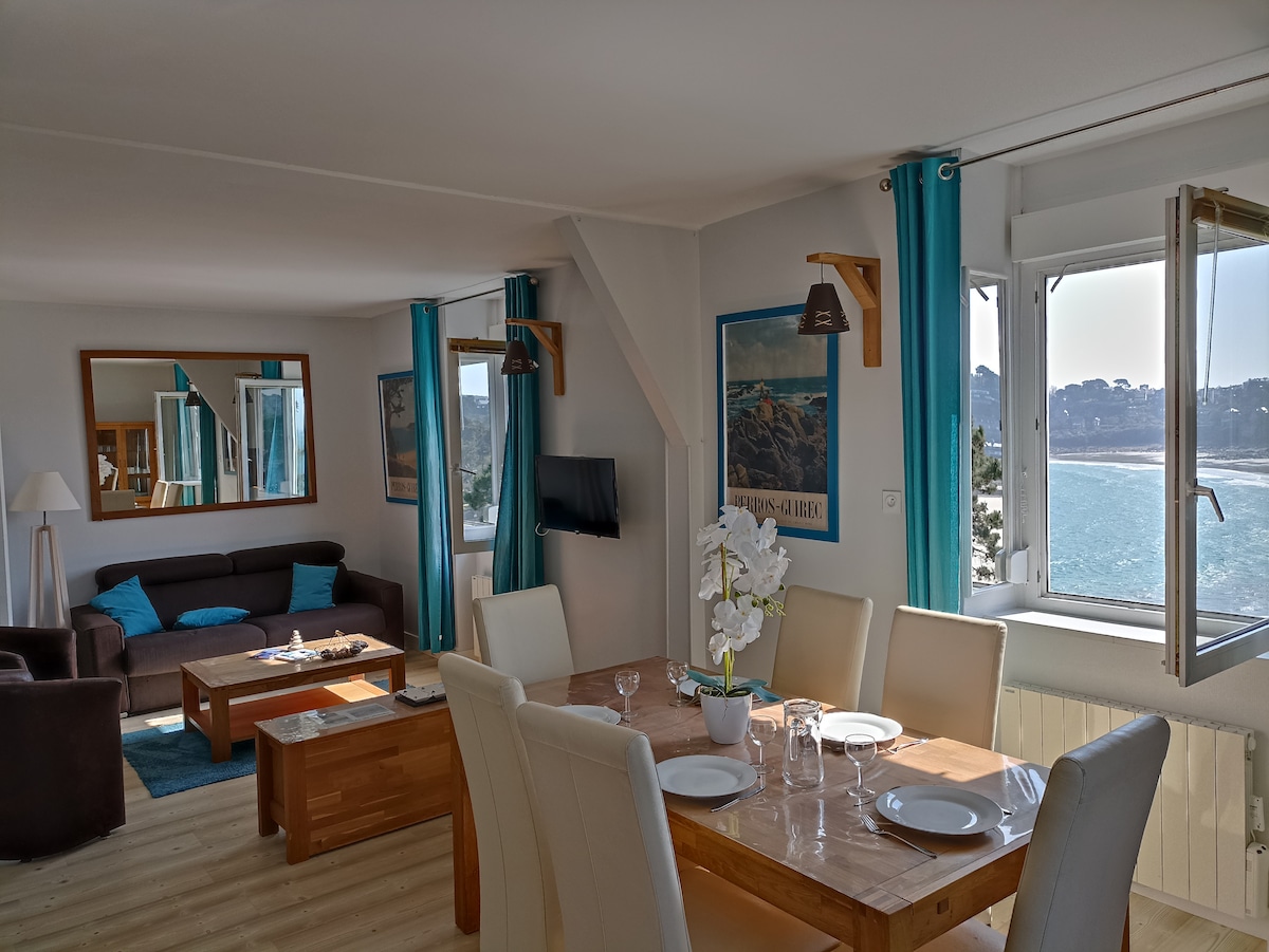 A living area with large windows offers scenic ocean views. A dark brown sofa is paired with a stylish wooden coffee table. A dining table with white chairs is set for a meal, complemented by decorative elements like a flower arrangement and wall art.