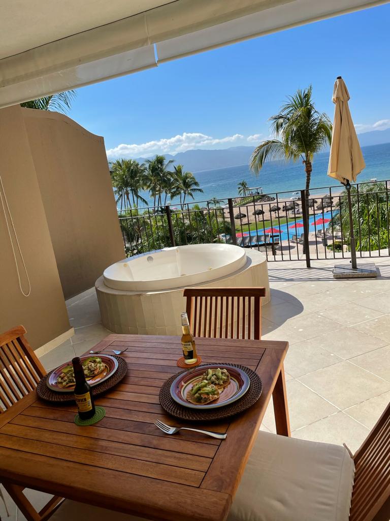 A private balcony features a wooden dining table set for two, adorned with plates of food and beverages. A hot tub is positioned nearby, with lush palm trees and beach views in the background, alongside a clear blue sky that enhances the outdoor dining experience.