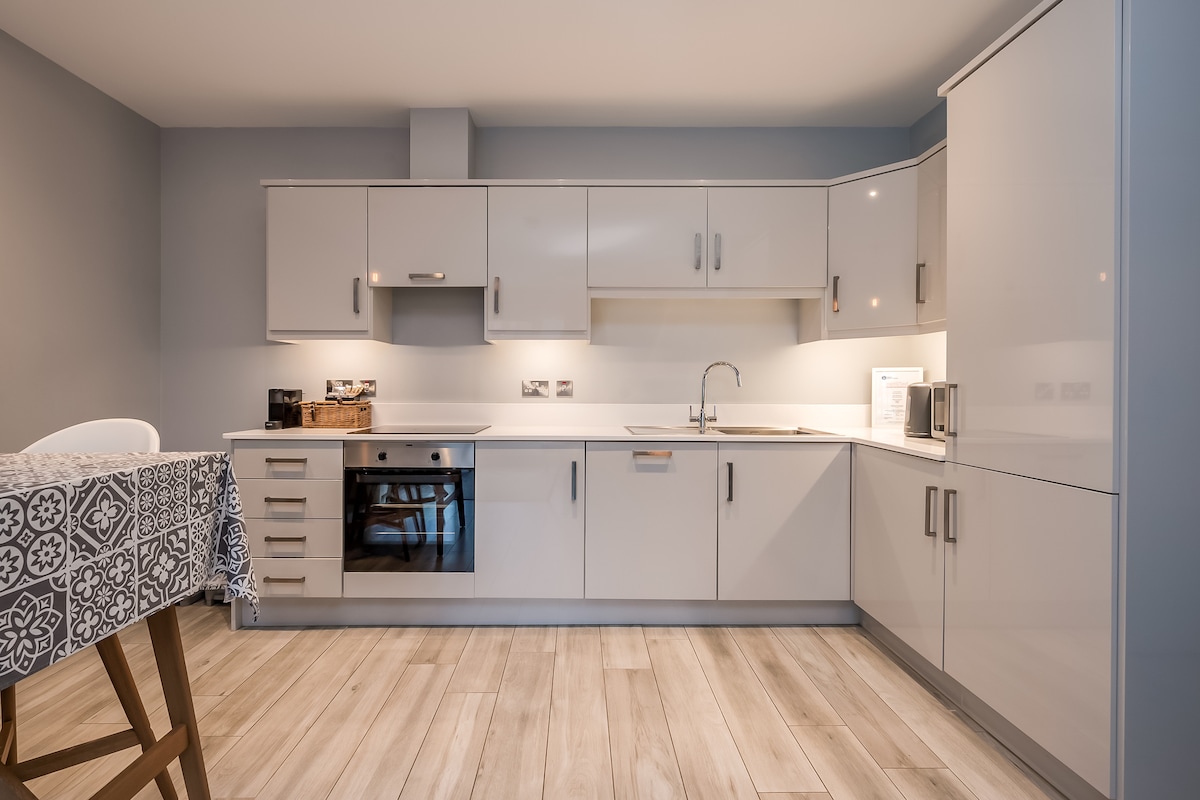 A well-equipped kitchen features sleek cabinetry and modern appliances, including a conventional oven and microwave. A small dining table is visible, accompanied by a set of chairs. Flooring in light wood tones enhances the clean, contemporary design.