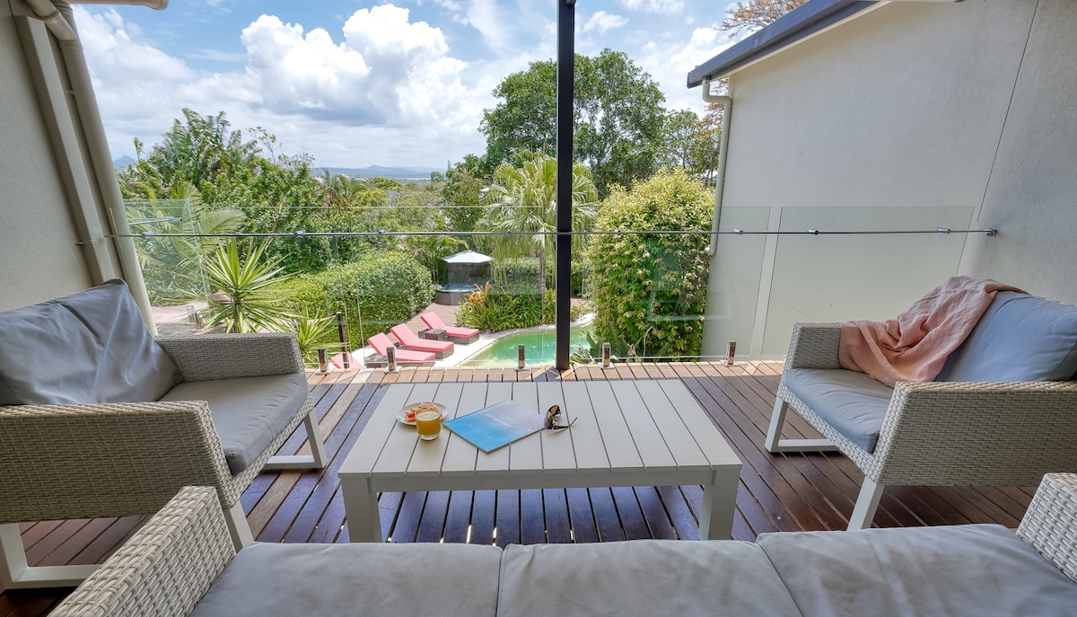 A covered timber deck features comfortable seating and a central coffee table. Through large glass panels, a view of the lush backyard with a lagoon-style pool is visible, surrounded by greenery and sun loungers, creating a relaxed outdoor atmosphere.