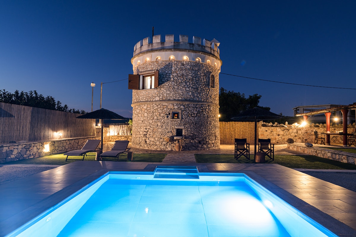 A stone tower stands elegantly against the night sky, complemented by a gently illuminated swimming pool. Two sun loungers are positioned nearby, and the surrounding area features stone walls and greenery, enhancing the privacy of the outdoor space.