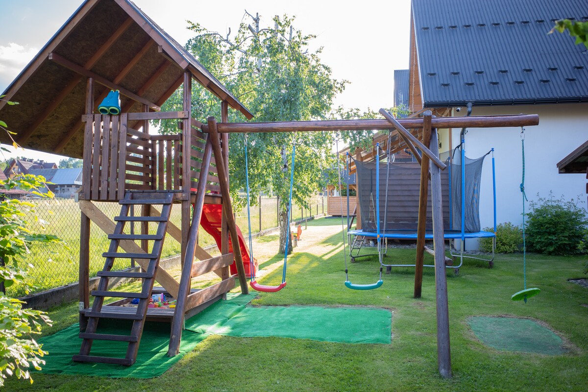 A play area is featured, equipped with a wooden play structure that includes a slide and climbing features. Two swings hang from a sturdy frame, while a trampoline is positioned nearby. Lush green grass surrounds the setup, creating a welcoming environment for outdoor play.