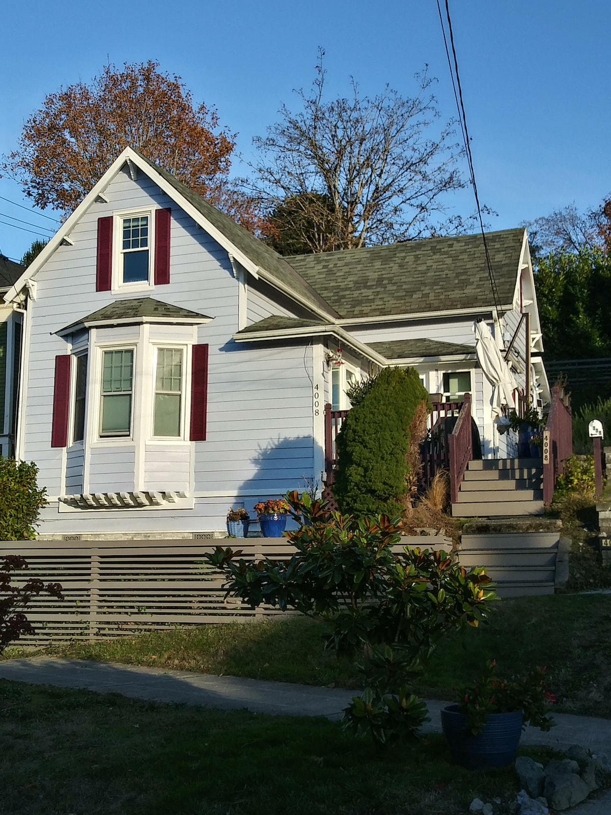 A charming early 20th-century house features a light blue exterior with deep red shutters. The front porch is framed by greenery, leading up to steep stairs. A manicured lawn and shrubs complement the welcoming entrance, illuminated by soft afternoon sunlight.