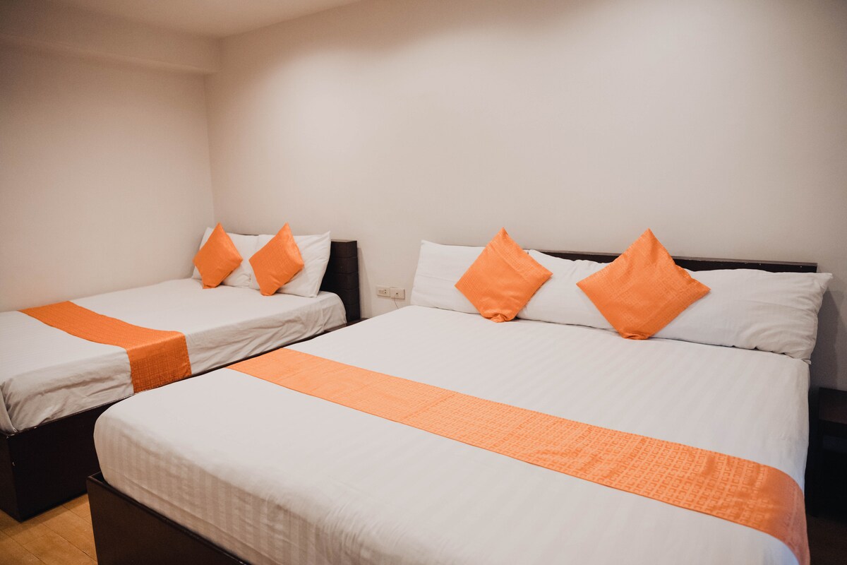 The room contains two beds, one queen and one king, both dressed in crisp white linens. Orange accent pillows are placed at the head of each bed. A simple wooden frame supports the bedding, and the space is illuminated by overhead lighting.